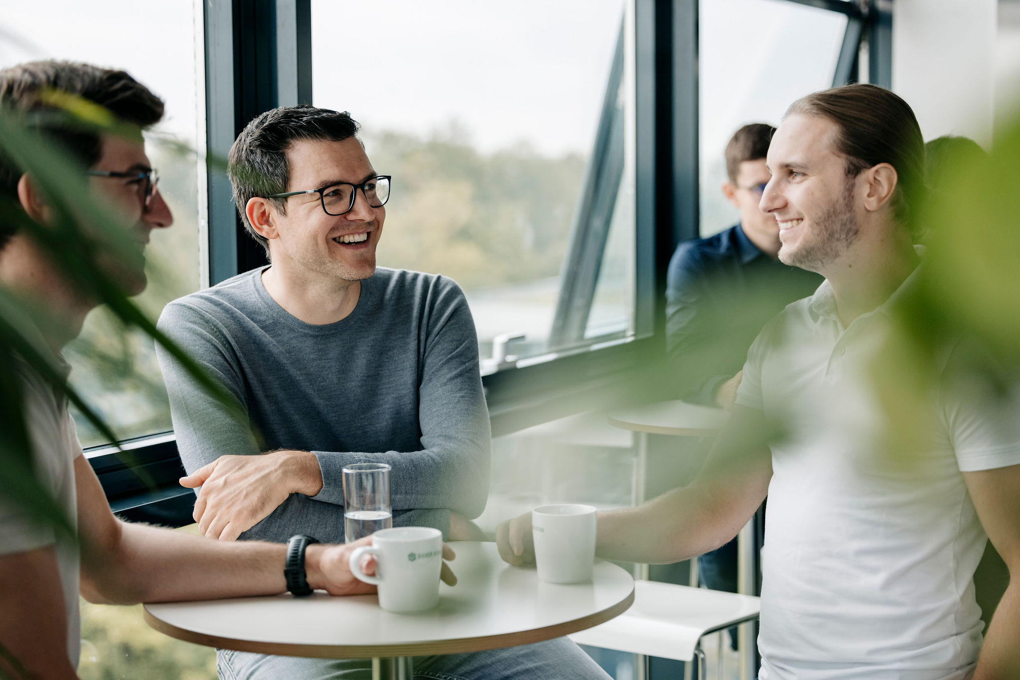 Gruppe an einem Stehtisch in einem Aufenthaltsbereich mit Fenstern im Hintergrund.